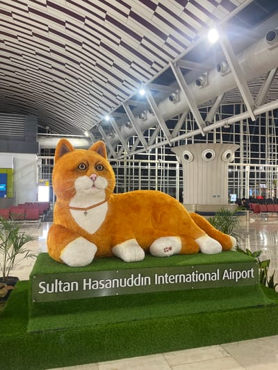 The image shows a large, orange plush cat statue with white paws and belly, wearing a red collar, lying on a green platform. Next to the statue is a sign that reads "Sultan Hasanuddin International Airport." The statue is located inside the airport terminal building, which is in Baji Mangngai, Indonesia. The building has a curved ceiling with patterned maroon and white lines and a metal framework. There are overhead air vents and lighting fixtures visible. In the background, there are airport seating, signage, and other people.