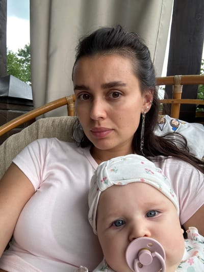 A mother and baby are posing for a selfie outdoors, likely on a covered porch or balcony in Sylva, Russia. The mother, with dark hair and brown eyes, is wearing a light pink t-shirt and a dangling earring. She holds the baby, who has blue eyes and is wearing a white hat with a floral pattern and a pink pacifier. The background shows a wooden railing, beige curtains, and greenery, suggesting an outdoor setting. The lighting is natural and appears to be daytime.
