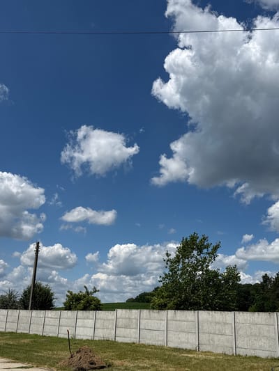 The media file shows a bright, daytime outdoor scene with a prominent blue sky filled with fluffy white and gray clouds. To the right, a tall, gray concrete fence runs horizontally, with sections demarcated by vertical lines. A strip of green grass runs along the base of the fence, and beyond that, a dense line of green trees forms a backdrop. In the foreground, to the left of the fence, a single dark power line runs diagonally upwards. Further to the left, a wooden pole with electrical components is visible. The scene appears calm and open, with no people or animals visible. The lighting suggests it is a sunny day, likely mid-morning or afternoon. The location is identified as Wysiedle, Poland.