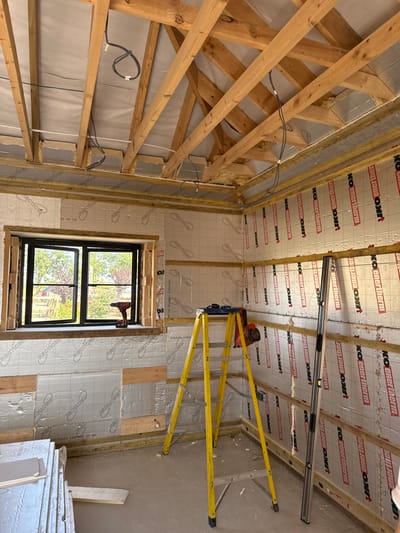 The image displays the interior of a room under construction in Skipsea, United Kingdom. The walls are partially insulated with silver-faced rigid insulation boards, identifiable as "Recticel" on the left wall and "IKO enertherm" on the right, secured by horizontal wooden battens. The ceiling structure consists of exposed wooden rafters and joists, with white insulating material visible between them, and black electrical wiring loosely run across. A black-framed casement window on the left wall is open, revealing a rural outdoor scene comprising green foliage, trees, and a wooden fence. A red and black cordless drill rests on the windowsill. A yellow A-frame stepladder is positioned centrally, with a red and black circular saw on one of its steps. A long, silver spirit level leans vertically against the right wall. Stacks of uninstalled grey and white construction panels are located on the unfinished concrete floor in the lower-left foreground. No individuals are present in the scene.