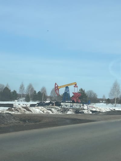 The scene is set outdoors in Ustinovo, Russia, under clear, light blue sky conditions consistent with late winter or early spring. A stationary oil pumpjack is the prominent feature in the midground. This pumpjack consists of a yellow walking beam, a red horsehead counterweight, and a blue supporting structure. The inscription "ОДИН-8-2-21" is visible on the yellow beam. The ground surrounding the pumpjack consists of melting snow, dark earth, and minimal vegetation, enclosed by a low, basic perimeter. In the background, a mixed tree line of bare deciduous trees and evergreen conifers stretches horizontally, with faint outlines of distant structures discernible through the trees. The foreground features a dark asphalt road, angled from the lower-left to the right, bordered by irregular mounds of melting snow and dark soil. A small, dark, indistinct aerial object is visible high in the clear sky, situated in the upper-left quadrant of the frame. No individuals or active movements are observed.