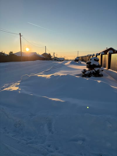 The image displays a snow-covered residential street in Stary Chultem, Russia, at sunrise or sunset. The sun is low on the left horizon, partially obscured by a distant roof, casting warm, golden light across extensive snowdrifts. These drifts line both sides of the unpaved or snow-covered road, where faint vehicle tire tracks are visible in the snow on the left. Utility poles and overhead lines run along the left side, adjacent to fences and residential buildings. On the right, a long corrugated metal fence with brick pillars parallels the street, interspersed with several small, snow-laden evergreen bushes. Distant houses line both sides of the road. A single aircraft contrail marks the clear blue sky. A small, circular green lens flare is visible in the lower right quadrant of the frame. No individuals are present.