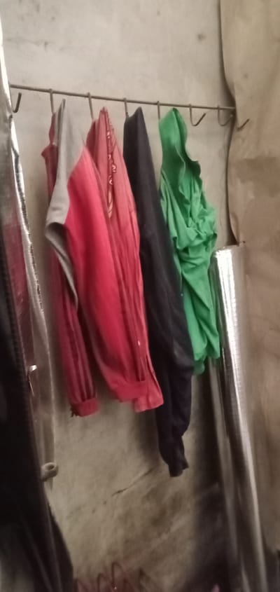 In the city of Sumput, Indonesia, several garments are hanging on a metal rod. From left to right, these include a metallic object that could be a rolled-up blanket or tarp, followed by a red and grey jacket, a predominantly red jacket with some patterned details, a dark navy jacket, and a bright green hooded jacket. The background is a rough, textured wall. To the right of the hanging clothes, a large, reflective cylindrical object is visible, likely another rolled-up material. The scene appears to be an indoor storage area or makeshift clothing line.