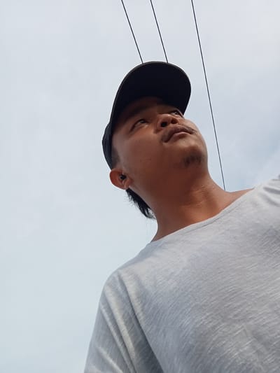 A close-up, low-angle shot shows a man wearing a black baseball cap and a white t-shirt, looking upwards against a cloudy sky with power lines overhead. He has a mustache and a small goatee. An earbud is visible in his left ear. The image appears to have been taken outdoors, possibly in Teluk Kenidai, Indonesia, based on the context provided, although no specific landmarks or identifying features of the location are visible. The lighting suggests it is daytime.