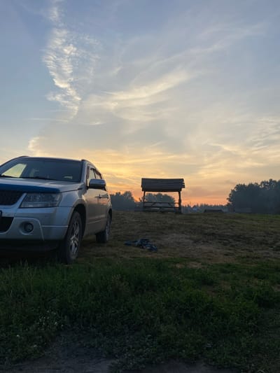 In Peshcherka, Russia, a silver SUV is parked on a grassy field under a sky with wispy clouds and the soft glow of sunset. In the background, a wooden gazebo with a weathered roof stands on a slight incline, with a row of trees forming a dark silhouette against the orange and yellow horizon. Two pairs of dark sandals lie discarded on the dry grass near the gazebo. The foreground is dominated by lush green grass, with the lower portion of the SUV visible on the left. The scene depicts a tranquil, rural outdoor setting during twilight.