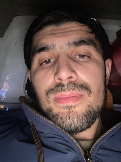 A man with brown eyes and a trimmed beard is centered in the frame. He wears a black beanie and a blue zip-up jacket with a brown collar peeking out. The image appears to be taken indoors, possibly in a car or a room with dull lighting and gray or white surfaces visible around the man. He is facing the camera with a slight upward tilt of his head.