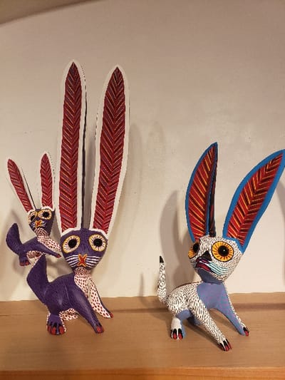 Three vibrantly painted wooden alebrijes resembling bird-like creatures are displayed. The top figure exhibits a white body speckled with black dots, striking yellow eyes, and wings colored with red and blue patterns. Below, two purple-bodied figures follow, with wings displaying red and white patterns. The alebrijes are mounted on a white wall with a tan wooden frame on the right, creating a visually captivating display of Mexican folk art.