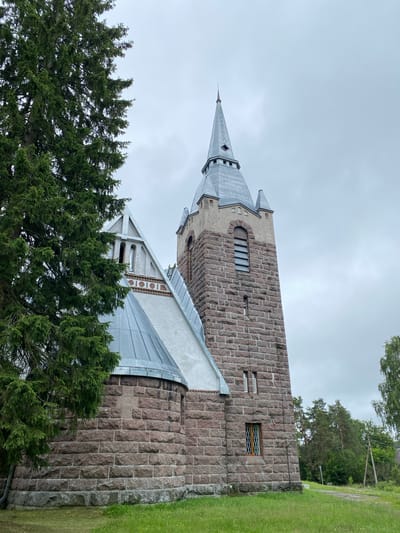 The image shows a church with a tall stone tower and a conical roof, partially obscured by a large evergreen tree on the left. The tower is constructed from rough-hewn, reddish-brown stones. It has several windows, including a tall, narrow one with horizontal slats near the top and a smaller, diamond-paned window lower down. The church has a grey, metal roof that slopes downwards. The sky is overcast with grey clouds. Green grass covers the ground in the foreground, and more trees are visible in the background. The scene appears to be in a rural or semi-rural setting.