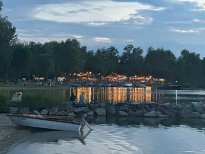 In Rimsting, Germany, a twilight scene unfolds by a lake. A small white rowboat with the registration TS-10632 is beached on the gravelly shore in the foreground. The lake's surface reflects the lights of a distant shoreline, where numerous people are gathered, enjoying what appears to be an outdoor event or festival. String lights crisscross the treeline, illuminating food stalls and tents. In the middle ground, a few individuals are seated on a rocky breakwater, facing the water. To the left, two children are sitting on the grass near the water's edge. The sky above is a blend of pale blue and gray clouds, suggesting the transition from day to night. The atmosphere is calm and festive.