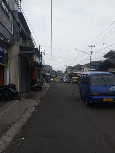 The image depicts a street scene in the city of Houaythat, Laos, under an overcast sky.
In the foreground on the right, a blue minivan, possibly a Suzuki Carry model, is parked facing away from the viewer. Its front license plate, "F 1904 YS" with "03-27" below, is visible. The words "NGE AYAP" are written in white letters on the upper part of the windshield, along with a "Family Gathering" sticker on the lower right.
On the left side of the street, a concrete sidewalk runs parallel to the asphalt road. Several motorcycles are parked on this sidewalk, with some partially covered by black plastic bags. Adjacent to the sidewalk is the lower portion of a multi-story building. An open doorway on the ground floor reveals a dark interior where a person, wearing a dark shirt, is seated. A blue banner with the partial text "UTER" is visible on the building's left side.
The asphalt road extends into the midground, where additional vehicles are present. These include multiple yellow minivans, white cars or vans, and more parked motorcycles lining the left side of the street. Several pedestrians are visible further down the left sidewalk. Buildings with varying architectural styles and storefronts line both sides of the road, many featuring awnings or overhangs. Visible signage includes a circular "PIZZA" sign and a larger sign displaying "Pemasaran."
In the background, the street continues, appearing to ascend slightly. The sky above is uniformly light grey and overcast. A dense network of utility wires, connected to multiple wooden and metal poles, crisscrosses overhead. A streetlight is also visible, along with a rooftop antenna on one of the buildings. The road surface shows signs of wear, and minor litter, including a red wrapper, is present on the asphalt near the curb in the foreground.