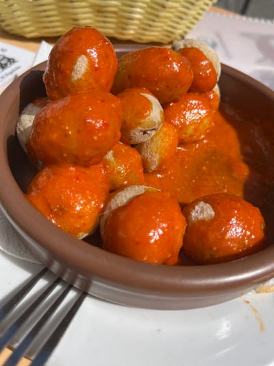 The image shows a brown ceramic bowl filled with papas arrugadas (wrinkled potatoes) covered in a bright orange mojo rojo sauce. The bowl is sitting on a white plate with a silver fork to the left. The potatoes are small, round, and appear to be boiled. The sauce is thick and coats the potatoes completely. The bowl is slightly tilted, revealing the contents. This meal appears to be a traditional Canarian dish, served in a restaurant in the municipality of Agüimes, located on the island of Gran Canaria in the Canary Islands, Spain. The geographic coordinates 27.907, -15.447 correspond to the municipality of Agüimes.