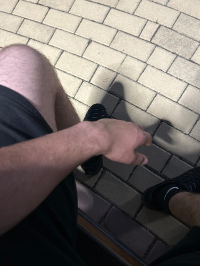 An overhead, low-light image depicts a person seated, looking down at their legs, right arm, and the ground in Dzhebel, Bulgaria. The individual's bare, hairy thighs and lower legs are visible, covered by black shorts. Black athletic shoes are worn on both feet, with a black sock featuring a white Nike swoosh visible on the right ankle. The person's bare right forearm and hand are extended forward, positioned over the paved surface. The ground consists of light-colored, rectangular paving stones laid in a staggered pattern with dark grout lines. Shadows, likely cast by the individual or an overhead light source, are visible on the paving. A dark, horizontal structure, possibly the edge of a bench or step, is present at the bottom of the frame.