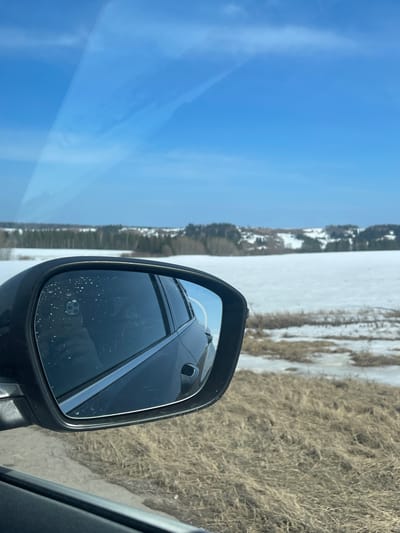 The image shows a view from inside a car, with the side mirror prominently featured. The mirror reflects the rear of the car and a portion of the landscape. Outside the car, there is a clear blue sky on one side, and a landscape of dry grass and patches of snow on the other. A line of trees and buildings can be seen in the distance. The photo was not taken in Kueda, Russia.