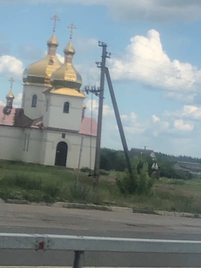 A white church with a golden dome is visible in the background. The church has two prominent golden domes topped with crosses, and a smaller golden dome in the distance. The building has a red roof and arched windows and doorways. A utility pole stands in front of the church. In the middle ground, there is a line of trees and some houses with red roofs. The foreground shows a road and a guardrail. The sky is blue with scattered white clouds. The scene is set in Rivnopillia, Ukraine.