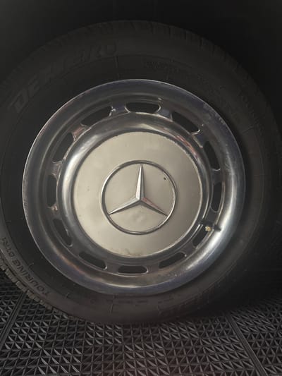 A close-up, eye-level forensic photograph depicts the passenger-side front wheel of a vehicle, possibly a vintage Mercedes-Benz, parked on a dark, interlocking, textured floor tile surface within an interior setting in Miami Shores, United States of America. The tire, a "DEXTERO TOURING DTR1," is dark black with visible sidewall text. A prominent, reflective chrome hubcap is affixed to the wheel, featuring a cream-colored circular center emblazoned with a silver Mercedes-Benz three-pointed star logo. Several elongated oval cutouts are visible around the perimeter of the hubcap. A brass-colored valve stem protrudes from the hubcap's lower right quadrant. The hubcap exhibits minor scuffs and surface dirt. No individuals, actions, or interactions are visible.