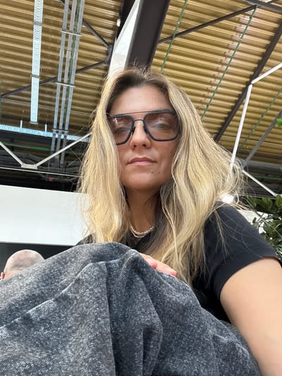 A woman with blonde hair and glasses is looking directly at the camera. She is wearing a black t-shirt and a pearl necklace. She is sitting down, and part of her body is covered by a dark grey blanket. In the background, there are industrial-looking structures and a yellow ceiling. To the left, the back of a person's head is visible. The scene appears to be indoors, possibly in a public space or an industrial area. The image is a close-up, with the woman's face and upper body being the primary focus. The location is specified as Perepechino, Russia.