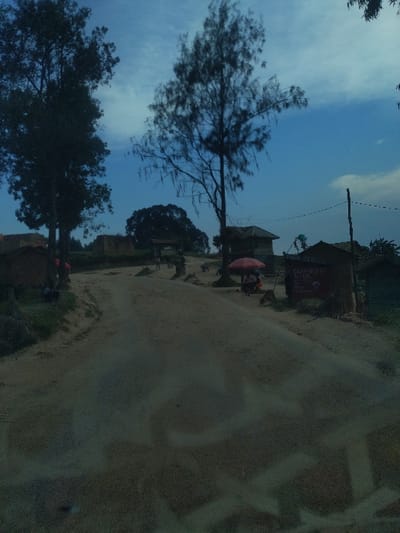 The image depicts an unpaved dirt road ascending gently through a rural or semi-urban area in Alimbongo, Democratic Republic of the Congo, under an overcast sky with visible blue patches. In the foreground and mid-ground, the road exhibits tire tracks or wear patterns. On the left, two large, dark-leafed trees frame the scene, with several small, simple structures, possibly made of mud or brick, visible behind them. Faint human figures are discernible near these structures. To the right of the road, a tall, sparsely-leafed tree stands. Further right, a small building features a prominent black sign displaying "CASH-POINT" and "AIRTEL MONEY," indicating a mobile money service point. A red market umbrella is positioned in front of this building, with an indistinct human figure visible beneath it, suggesting a vendor. A utility pole with a horizontal wire is present on the far right. Additional small structures and indistinct human figures are scattered in the distant background along the road.