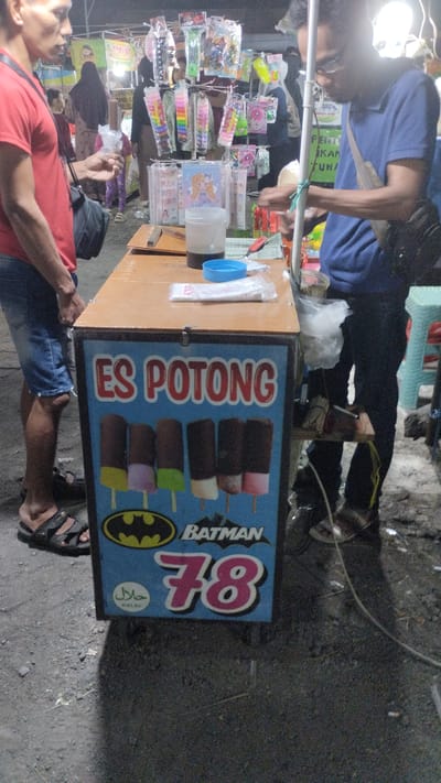 The image depicts a street vendor selling ice cream in Sambungrejo, Indonesia. The central subject is an ice cream cart, a freezer chest with a wooden top, adorned with a vibrant blue graphic advertising "ES POTONG" (cut ice cream) and featuring images of various ice cream bars, the Batman logo, and the number "78". A "HALAL" certification is also visible.
In the foreground, a man wearing a red t-shirt and denim shorts stands on the left, holding a small item in his hand. On the right, another person, wearing a blue t-shirt and dark pants, is partially visible, appearing to interact with the cart or serve a customer. The background reveals a bustling market or roadside stall setting with other vendors and displays of merchandise, including colorful toys and accessories. The lighting suggests it might be evening or night, given the artificial lights illuminating the scene. The overall atmosphere appears lively, characteristic of a local market. Visible text includes "ES POTONG", "HALAL", "BATMAN", "78", and some partial Indonesian text on signage in the background, such as "JASUK MANG: 6, AR: 11", "PENT", "KAN", and "TUN".