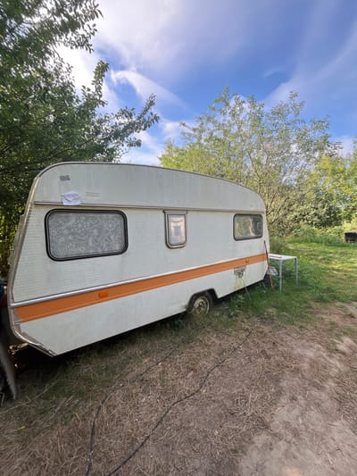 The scene is located in an outdoor, overgrown area within Sint-Pieters-Leeuw, Belgium, under a clear blue sky with scattered cirrus clouds. The primary subject is a weathered, off-white single-axle caravan with a distinctive horizontal orange stripe running along its lower side. The caravan's exterior shows dirt accumulation and minor surface irregularities. An irregular white patch, possibly a piece of paper or tape, is affixed to the upper front left panel of the caravan. Three windows are visible on the side: a large, rectangular window at the front with a dark frame and an obscured interior covered by a white, patterned curtain or decal; a smaller, vertical rectangular window in the middle; and another rectangular window towards the rear. An amber side marker light is present on the orange stripe, and a rectangular vent is visible below the central window. The caravan rests on an uneven ground composed of patchy, dry grass and exposed soil. A black electrical cord extends from near the front underside of the caravan, trailing across the ground. To the right of the caravan, a small, light-colored rectangular table with metal legs stands partially in the grass. Leaning against the caravan next to the table is a long, thin, brown object resembling a wooden stick or cane. The caravan is surrounded by dense green foliage, including trees and bushes, which form a backdrop and partially obscure the view. No individuals are present, and no active interactions or actions are depicted within the frame.