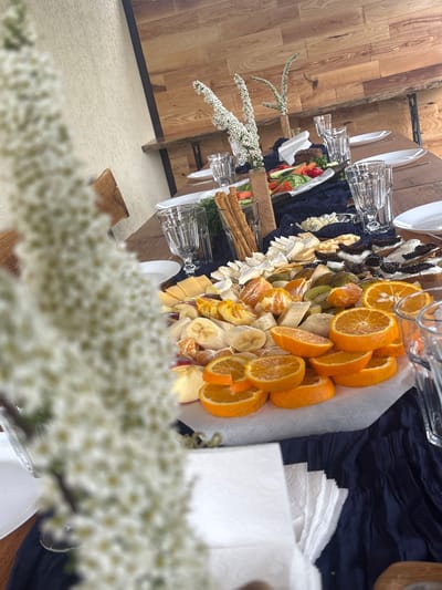 The image shows a long table set with plates, glasses, and a variety of food items, including sliced oranges, bananas, cheeses, and vegetables. The table is covered with a dark blue cloth under the food items, and there are white napkins and small floral arrangements as decorations. The table is made of wood, and the floor surrounding it is a light beige color. The location is likely indoors, possibly in a dining area, and the blurry foreground suggests the photo was taken through or near a floral arrangement, potentially indicating a special occasion or celebration. There is no evidence to confirm that the location is Lahojsk selsaviet, Belarus.