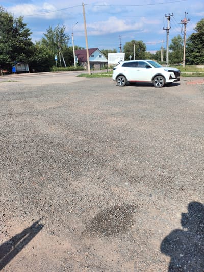 A white SUV is parked on a gravel lot in Pokhvistnevo, Russia. The SUV has red accents on its side. In the background, there are trees, a bus stop, and a building with a brown roof. The sky is blue with scattered clouds. The image is taken from a low angle, with the shadows of two people visible in the foreground, suggesting the photographer is one of them.