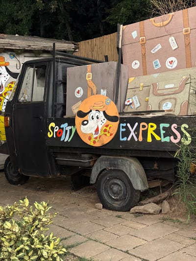 A black three-wheeled vehicle, resembling an auto-rickshaw, is parked on a paved surface. The vehicle's side panel displays colorful painted lettering spelling out "SPOTTY EXPRESS". To the left of the lettering, a circular orange panel features a cartoon drawing of a Dalmatian dog wearing a hat and a bow tie, with pizza slices near its mouth. The vehicle is loaded with large, brown, suitcase-like structures, painted with details of straps, buckles, and travel stickers. One sticker on the largest suitcase reads "ORLANDO". The background includes a wooden fence and trees, suggesting an outdoor setting. The scene is located in the city of Cavriglia, Italy, likely within an area decorated to evoke a whimsical, travel-themed atmosphere.