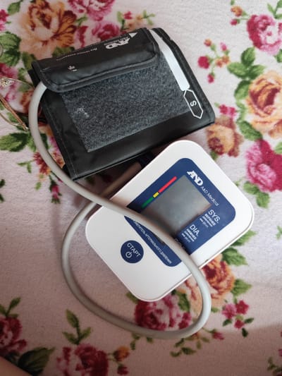 An A&D Medical blood pressure monitor with a cuff is placed on a floral-patterned cloth. The cuff is dark grey with a light grey border and a velcro closure. A white tube connects the cuff to the white digital display unit. The display unit has a dark blue screen with red, yellow, and green indicator bars. The button for starting the device is labeled "СТАРТ" in Russian, meaning "START". The device is positioned on the right side of the image, with the cuff extending to the left. A pair of eyeglasses with gold frames is partially visible on the left side of the image. The background is a pink, white, and green floral fabric. The image appears to be taken indoors. There is no information in the image to indicate the location in Yenakiieve, Ukraine.