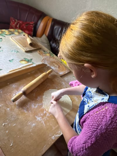 A young girl with blonde hair is shown from behind, as she prepares dough on a wooden surface. She is wearing a blue and white floral apron over a pink knitted sweater. A rolling pin is visible on the table in front of her, along with a flattened piece of dough. In the background, a portion of a brown leather couch with red and yellow accents can be seen. The overall setting appears to be a domestic kitchen or dining area, possibly in Valday, Russia, given the context.