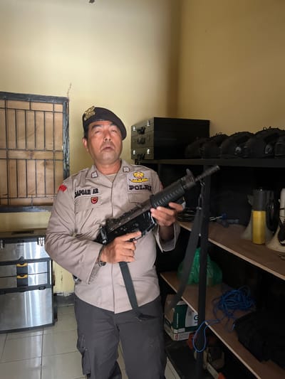 A man in a tan police uniform with a black beret holds a rifle. The man has a dark complexion, short dark hair, and is looking forward with a neutral expression. He is wearing a name tag that reads "SAPOAN AR" and a badge on his uniform. The man is standing in a room with yellow walls. To his left is a metal cabinet with drawers, and a window with metal bars. To his right are shelves filled with black helmets, a yellow canister, and other objects. The rifle he is holding is black and appears to be an assault rifle. The scene suggests a police storage or armory area. The location is identified as Tanakawu, Indonesia.