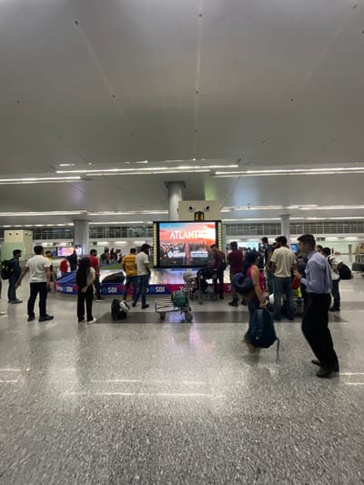 The image depicts a busy scene inside a modern airport terminal, likely at Raipur Khurd, India. A large digital display screen advertises "ATLANTIS GRAND" with an image of a cityscape. Below the screen, a baggage carousel is visible, with several people standing around it, presumably waiting for their luggage. A variety of individuals, dressed in casual attire, are present in the frame, some standing, some walking, and others interacting. Luggage, including backpacks and rolling suitcases, is scattered throughout the area. The terminal has a high ceiling with strip lighting and large pillars. The floor is made of polished grey speckled material. The overall atmosphere suggests a typical travel hub with people in transit.