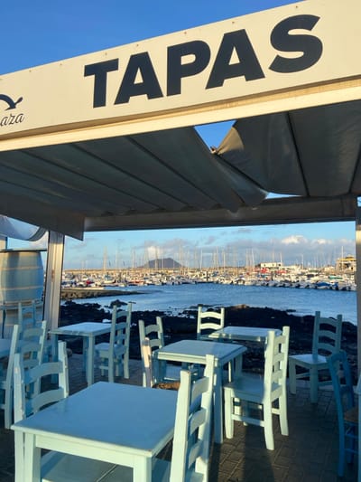 The image is taken from inside a restaurant in Corralejo, Spain, specifically at the coordinates 28.741 latitude and -13.866 longitude. The restaurant's awning, which is made of canvas material, is partially open and shows the harbor with boats and a distant island, while the blue sky is visible in the upper left corner of the image. The awning is marked with the word "TAPAS" in large, bold, black letters. The image focuses on the restaurant tables and chairs which are made of light blue wood. They are positioned on a black sand floor, with a view of the harbor. There are empty tables and chairs, suggesting that no one is currently using the seating area.