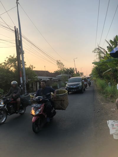 A street scene in Glagah Arum, Indonesia, at dusk. Two men are riding motorcycles on a paved road. The man on the left, wearing a helmet, is on a red motorcycle and is carrying a woven basket. The man on the right, wearing a blue shirt, is on a red scooter and is also carrying a large woven basket. A black SUV is driving in the same direction behind them, followed by another motorcycle with two riders. To the right of the road, tall grass and banana trees are visible. Utility poles with numerous wires line the left side of the road, where houses with tile roofs can be seen. The sky is a pale orange and yellow, indicating the sun is setting. Bags of what appears to be cement or fertilizer are stacked on the side of the road in the foreground on the right.