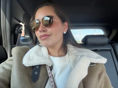 A person with brown hair and round sunglasses poses inside a car. The individual wears a light-colored, fuzzy collar jacket over a white sweater. The car's interior features dark leather seats and trim, with a headrest visible in the background. A portion of the car window shows a blurred outdoor scene. The photograph appears to be taken from a slightly elevated angle, with the camera capturing the person and interior details of the vehicle.