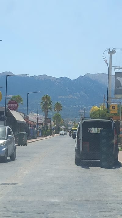 A street scene in Tigaki, Greece, shows a paved road lined with shops and palm trees. Mountains form the backdrop. The sky is clear blue. On the left, a small silver car is parked. Further down the street, several shops display signs, including one for "BROTHERS". A dark van is in the foreground, in the right lane of the road, facing away from the camera. Other vehicles, including a car and motorcycles, are visible further down the street. The overall scene suggests a sunny day in a Mediterranean town.