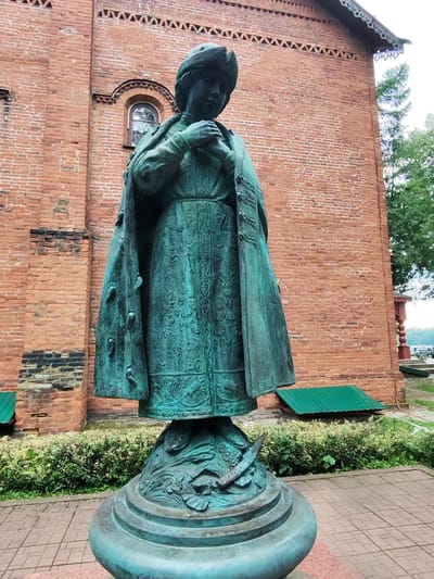 A full-body bronze statue stands on a stone pedestal in front of a red brick building. The statue depicts a woman wearing a fur-lined hat and a long coat over a patterned dress. She is holding her hands clasped in front of her. The building behind her has arched windows and decorative brickwork near the roofline. Green bushes and trees are visible to the right of the statue, and a paved walkway is in the foreground. The image is taken from a low angle, looking up at the statue. The location is identified as Chelganovo, Russia, based on the provided information.