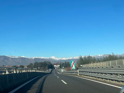 This media file captures a wide-angle view of an empty highway (autostrada) near Caselle Torinese, Italy, under a clear, bright blue sky. In the background, a majestic range of snow-capped mountains stretches across the horizon, adding a dramatic natural element to the scene.
The highway, made of asphalt, features clear white lane markings and gently curves towards the right as it extends into the distance. Metal guardrails line both sides of the road, with an additional concrete barrier topped with more guardrails visible on the left. On the right side, a sound barrier constructed from vertical panels is present, behind which a line of evergreen trees can be seen. Further in the mid-ground, beyond the highway, some buildings and structures are visible amidst more trees.
A prominent green and white road sign is situated on the right side of the road. This sign, characteristic of Italian motorways, displays a white letter 'A' with a diagonal red line through it, indicating the end of a motorway section or an exit.
The scene is bathed in natural daylight, suggesting a clear and sunny day, likely in the morning or early afternoon, given the crisp shadows and overall illumination. The weather appears fair with excellent visibility extending to the distant mountain peaks. No people are visible in the image, emphasizing the open and expansive feel of the road. The setting evokes a sense of serene travel through a scenic, mountainous landscape.