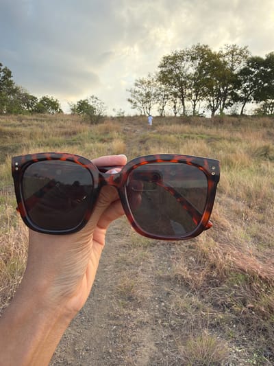 A person's hand holds a pair of tortoiseshell sunglasses in the foreground, obscuring the lower part of the view. The sunglasses have dark lenses that reflect the sky and surrounding environment. The hand is positioned in the lower left of the frame. Behind the sunglasses, a dirt path winds uphill through dry, golden grass and scattered trees. In the distance, a lone figure in a white shirt and shorts walks up the path. The sky above is overcast with patches of sunlight. The scene appears to be an outdoor location, possibly a hillside or park in Kubutambahan, Indonesia.