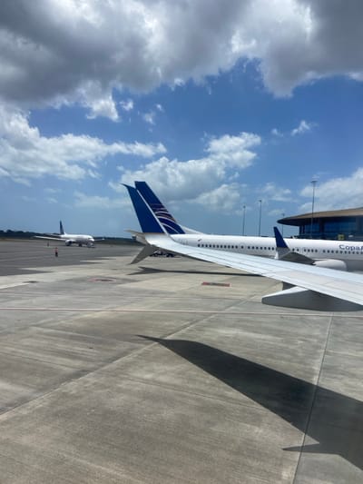 The image displays an airport apron scene at Tocumen International Airport in Tocumen, Panama, under a partly cloudy daytime sky. The perspective is from within an aircraft, with its white wing and a section of the fuselage visible in the lower right, casting a distinct shadow on the grey concrete tarmac.

In the mid-ground, a white Copa Airlines passenger jet is stationary, identified by its blue and white vertical stabilizer and "CopaA" lettering on the fuselage. Immediately behind this aircraft, a segment of an airport terminal building with a dark-colored roof and large windows is visible, flanked by tall floodlight poles.

Further left on the tarmac, a second Copa Airlines aircraft is parked. Near this distant aircraft, two individuals wearing high-visibility vests are present on the concrete surface, along with an unidentified piece of ground support equipment. The horizon in the far distance features a faint line of green vegetation. The sky exhibits patches of clear blue interspersed with white and grey cumulus clouds.