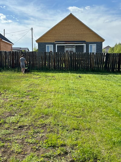 The image captures a daytime scene in Kuda, Russia, under a clear blue sky with scattered white clouds. The foreground consists of an expanse of green grass mixed with areas of bare soil. In the midground, a male figure, observed from the back, is seen wearing a grey short-sleeved shirt and grey shorts, pushing a green and black lawnmower across the grassy area. He is positioned in front of a dark brown wooden fence constructed from vertically oriented, irregularly shaped planks.
Beyond the fence, a residential house is visible, featuring a dark grey brick lower section and a prominent light brown wooden gable roof. This house has two white-framed windows and a central white-framed sliding door on its facade, partially covered by a dark, corrugated awning. To the left of this primary residence, another structure with reddish-brown brick siding and a dark roof is partially visible. Further in the left background, two utility poles with multiple overhead wires are present. Green foliage from trees is discernible in the far background on the right.