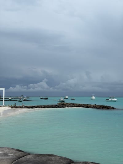The scene depicts a beach area in Fulidhoo, Maldives under an overcast sky with storm clouds looming. A rocky coastline separates the sandy beach from a calm, turquoise sea. Several boats are anchored in the water, varying in size and type. A white structure, likely part of a building or pier, is visible in the lower left corner. A large boulder is on the right edge of the frame. The location appears to be a typical tropical setting with elements of both natural beauty and human development. There are no people visible in the scene.