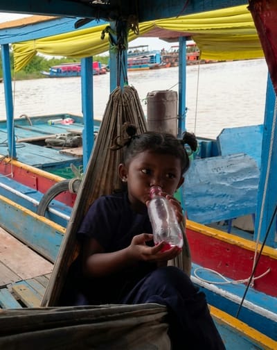 A young girl with pigtails is sitting in a hammock on a wooden boat. She is drinking from a plastic bottle with red liquid inside. The boat is on a river, and other boats are visible in the background. The scene appears to be in a rural area of Soutr Nikom, Cambodia, likely a floating village or market. The water is calm, and the sky is overcast. The boat is painted with bright colors, including blue, yellow, and red. The girl is wearing a dark blue shirt and pants. Her expression is neutral.