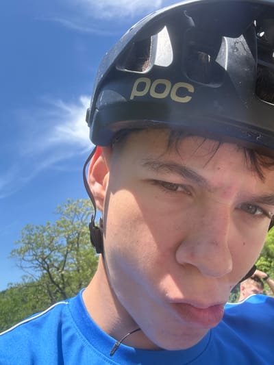 A close-up, tilted shot shows a person wearing a dark-colored POC bicycle helmet and a blue athletic shirt with white trim. The person's face fills much of the frame, revealing light skin, brown hair, brown eyes, and a slight blemish on their forehead. The background consists of a blue sky with wispy clouds and a tree with green leaves partially visible in the lower right corner. Another person is partially visible in the upper right corner of the frame. Based on the visible elements, there is no information to determine whether this photograph was taken in Donchovci, Bulgaria.