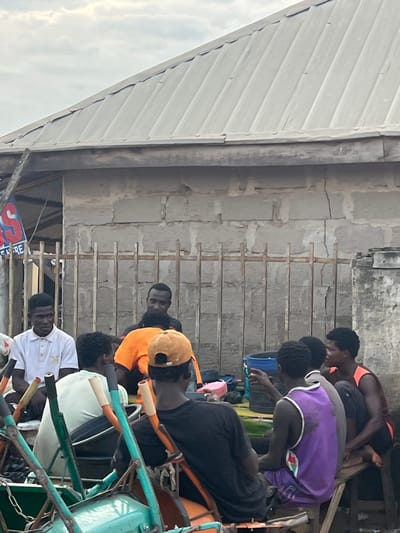 In Oko Amakom, Nigeria, a group of young men is gathered outdoors, seated around what appears to be a makeshift gathering or social space. The backdrop consists of a rough, concrete-block building with a corrugated metal roof, showing signs of weathering and cracking. A metal fence separates the immediate area from the building.
The men are varied in their attire. One man in the foreground wears a white polo shirt with a crest. Next to him, another man is dressed in an orange t-shirt. Further back, several other men are visible, including one wearing a purple tank top and another in a dark t-shirt. Some men are seated on wooden crates or benches.
In the foreground, parts of a green metal structure are visible, possibly a cart or a part of a transport vehicle, with visible chains and metal rods. A blue bucket and other small items are also present in the scene. The overall impression is of a casual outdoor gathering in a somewhat unrefined urban environment. The lighting suggests it is daytime, possibly overcast.