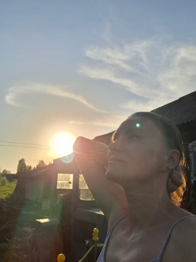 A low-angle, rotated outdoor photograph depicts a person against a bright sun and sky. The person, likely female, is fair-skinned with their head tilted back. Her hair appears to be blonde, catching the sunlight. Her left hand (as viewed) is raised behind her head. She is wearing a light-colored sleeveless top.
In the background, blurred shapes suggest buildings or structures, possibly rural or agricultural. There's vegetation present and a hint of yellow objects hanging off the structures. The sky is a gradient from light yellow to blue.
It is not possible to confirm the location of the city of Шешурга, Russia as there are no explicit landmarks or signs that would indicate a precise location. The scene depicts an individual relaxing outdoors, perhaps in a rural or semi-rural setting, during golden hour.