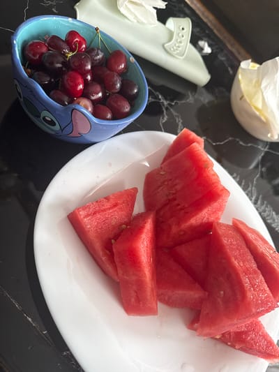 The image shows a table setting with a plate of watermelon slices and a small blue bowl of red grapes. Next to the grapes is a white cloth napkin. The table has a black, textured surface. No people are visible. The location is not identifiable as Momchilovtsi, Bulgaria based on the visual information.