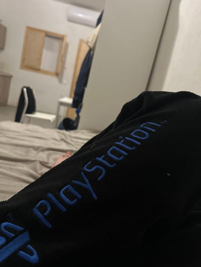 The image shows a close-up view of a black fuzzy garment with blue embroidered "PlayStation" lettering. The background is blurry, but includes a white wall with a small window, a chair, and a bed with beige sheets. The location in the city of Thira Municipal Unit, Greece, cannot be confirmed or denied based on the visual information provided.