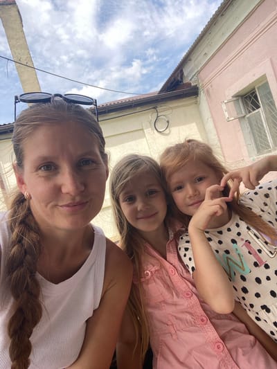 A low-angle, selfie-style photograph captures a woman and two young girls looking towards the camera, likely in Gladkovskaya, Russia. The image is oriented vertically with the sky and buildings at the top and the subjects' faces in the foreground.
The central subject is a woman with light brown hair, possibly with blonde highlights, styled in a braid over her left shoulder. She wears a white ribbed tank top and has sunglasses perched on her head. Her expression is a soft, content smile. Gold hoop earrings are visible on her ears.
Directly behind and slightly below the woman are two young girls. The girl on the left has blonde hair and wears a pink button-up shirt, showing a subtle smile. The girl on the right has red hair, a light-colored dress with black polka dots, and abstract letter-like shapes (appearing as "MN" or similar) on it. She is actively making a heart shape with her hands and also has a cheerful expression.
The background shows a blue sky with scattered white clouds, suggesting a clear day. Above the subjects, architectural elements are visible: a building with a pale pink facade and a window featuring security bars. Below this, an older structure with peeling paint, a brown drainpipe, and electrical wires is seen, indicative of an aged urban or semi-urban environment. The bright natural lighting suggests the photo was taken during daylight hours. All subjects appear happy and relaxed, suggesting a casual, joyful moment.