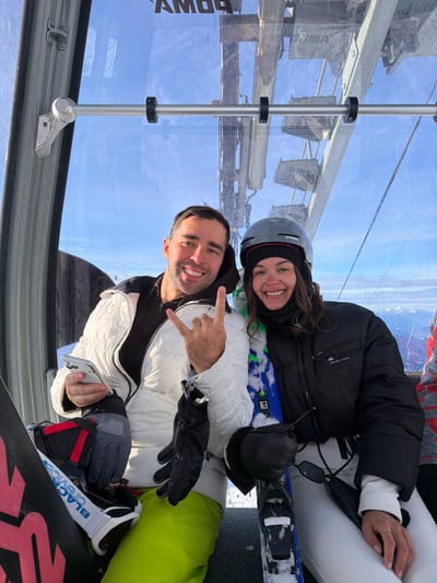 Two individuals, a man and a woman, are captured in a bright, outdoor setting, likely inside a ski lift or gondola cabin. Both are smiling broadly and looking directly at the camera, conveying a cheerful mood.
The man, positioned in the foreground on the left, has short dark hair and a clean-shaven face. He is wearing a white puffy ski jacket with black accents, black gloves, and light green ski pants. His left hand is raised, making a "rock on" gesture with his index and pinky fingers extended. In his right hand, he holds a silver or white smartphone with a light blue case. A black and red snowboard with white bindings and white-framed goggles is visible next to his leg. The text "BLACK CREW" and "K2" can be seen on the snowboard/bindings.
The woman, behind and slightly to the right of the man, has long dark hair peeking out from under a grey ski helmet with black accents. She is wearing a black knit neck warmer, a black puffy ski jacket (with a small triangular "PRADA" logo visible on the left sleeve), white ski pants, and black gloves. A pair of skis, featuring a blue and green pattern with black bindings, rests beside her. Dark sunglasses hang around her neck.
They are situated within a modern cabin with large, clear glass windows. Through these windows, a vibrant blue sky and distant snowy mountain peaks are visible, suggesting a high-altitude ski resort. A metal pole with black rings runs vertically inside the cabin. Reflected or partially visible text on the exterior of the lift mechanism or a nearby structure outside the window appears to spell "P O W Y", likely part of a word like "POWDER". The bright daylight and clear sky indicate sunny weather. The overall scene depicts a happy moment during a ski or snowboard outing.