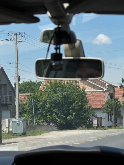 The view is from inside a vehicle, likely a car or truck, with the rearview mirror prominently in the foreground. The reflection in the mirror shows a person's face, partially obscured. Outside, the scene depicts a residential street. There is a building with a red tiled roof and beige brickwork visible. A large, green tree is situated next to the road. A power pole with lines runs along the street. The road has a yellow lane marking. The location appears to be in a rural area, possibly within Ruma Municipality, Serbia, given the architectural style and general environment. A trash container and a street sign are also visible next to the road.