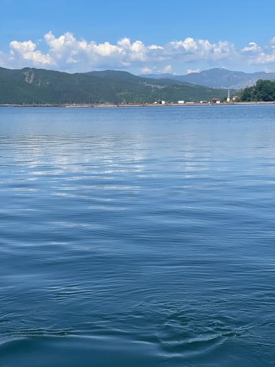 The image depicts a calm body of water, likely a lake or reservoir, with ripples on its surface. In the background, rolling green hills covered in trees rise against a bright blue sky dotted with fluffy white clouds. A low-lying structure, possibly a village or resort, is visible along the shoreline, with some buildings and a telecommunications tower. The water occupies the foreground and midground, reflecting the sky and clouds, creating a sense of depth and tranquility. The scene appears to be located in a natural, undeveloped area. There are no people or animals visible in the image. The overall impression is one of a peaceful natural landscape.